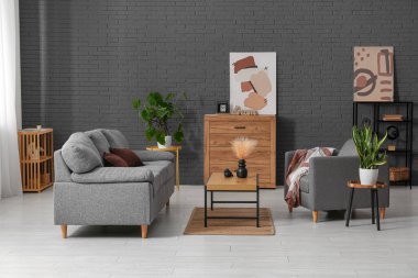 Beyaz tuğla duvarın yanında rahat gri kanepesi, koltuğu ve masası olan bir oturma odası.