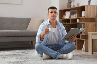 Oturma odasında yerde otururken elinde kahve fincanı olan yakışıklı bir genç.