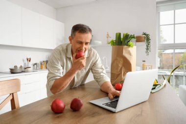 Elmalı olgun adam mutfaktaki masada dizüstü bilgisayar kullanıyor.