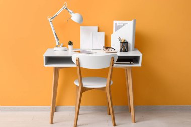 Tablet bilgisayarı, masa lambası ve turuncu duvarın yanındaki not defterleriyle rahat bir iş yeri.