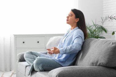Güzel genç bir kadın nefes egzersizi yapıyor ve evdeki kanepede meditasyon yapıyor.