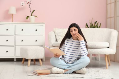 Genç, şaşkın bir kadın kitap okuyor ve şık bir oturma odasında yerde oturuyor.