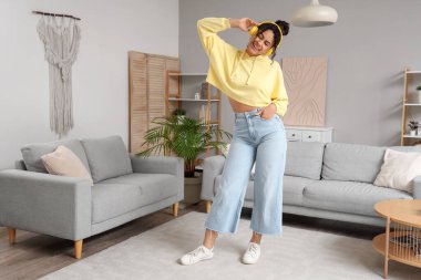 Güzel, genç, mutlu, Afro-Amerikan bir kadın kulaklıkla müzik dinliyor ve evde dans ediyor.