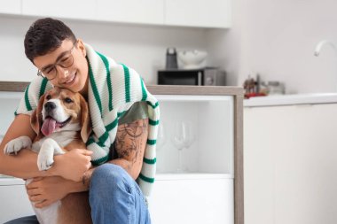 Gözlüklü genç adam mutfakta sevimli Beagle köpeğine sarılıyor.