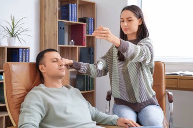 Cep saati olan bir kadın psikoloğu ofiste adamı hipnotize ediyor.