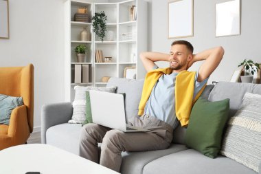 Evdeki kanepede dizüstü bilgisayarlı genç sakallı adam dinleniyor.