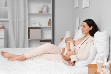 Pijamalı genç bir kadın bebeğini evde emziriyor.