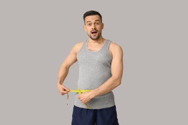 Yakışıklı, şoke olmuş bir adam. Gri arka planda belini ölçüyor. Kilo verme konsepti