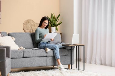 Oturma odasındaki kanepede doküman ve dizüstü bilgisayarla çalışan güzel genç bir kadın.