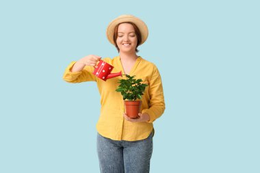 Hasır şapkalı gülümseyen kadın mavi arka planda çiçekleri suluyor.