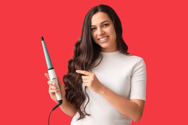 Beautiful young woman with curling iron on red background