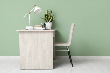 Ofiste yeşil duvarın yanında kırtasiye, lamba ve defter bulunan şık bir masa.