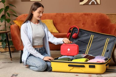 Evinde yerde oturan bavulu ve ilk yardım çantasıyla mutlu genç bir kadın. Seyahat kavramı