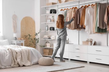 Pijamalı genç bir kadın aydınlık yatak odasındaki modern gardıroptan kıyafet seçiyor.