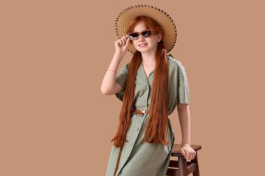 Şık giyinmiş hoş bir kızıl kız bej arka planda sandalyede oturuyor.