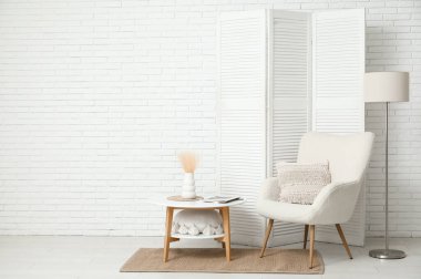 Modern odanın içinde katlanır ekran, koltuk, zemin lambası ve beyaz tuğla duvarın yanındaki masa.