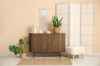 Bej duvarın yanında katlanmış perdeli, puf ve göğüs çekmeceleri olan modern bir oda.