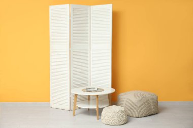 Sarı duvarın yanındaki katlanmış perdeli, puflu ve masalı odanın içi.