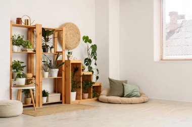 Odanın iç tarafındaki beyaz duvarın yanında raflar, puflar ve bitkiler var.