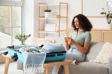 Genç Afro-Amerikalı kadın evdeki kanepede bavula seyahat aksesuarlarını yerleştiriyor.