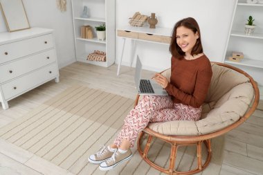 Dizüstü bilgisayarı olan genç bir kadın evinde koza sandalyesinde oturuyor.