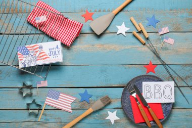 ABD bayrağından yapılmış çerçeve, 4 Temmuz 'da Mutlu Yıllar yazılı tebrik kartı, içinde barbekü malzemesi ve mavi ahşap arka planda barbekü aletleri olan kağıt levha. Bağımsızlık Günü kutlamaları