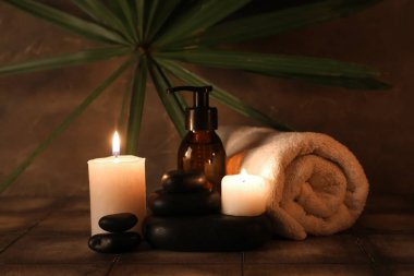 Bir şişe kozmetik ürünü, spa taşları, havlu ve yanan mumlar gri döşeme masasında, griler duvarın yanında, yakın plan.