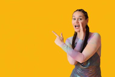Sarı arka planda bir şeye işaret eden rastalı saçlı dedikoducu genç kadın.