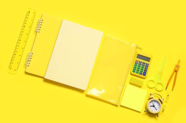 Alarm saati, hesap makinesi ve sarı zemin üzerinde farklı okul kırtasiyesi olan kompozisyon