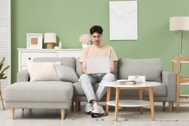 Gözlüklü genç adam evdeki gri koltukta dizüstü bilgisayar kullanıyor.