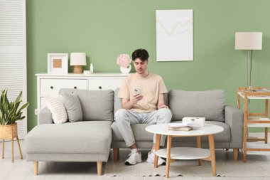 Evdeki gri koltukta cep telefonu kullanan genç adam.