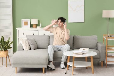 Kulaklıklı genç adam evdeki gri koltukta oturuyor.