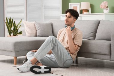 Kulaklıklı genç adam evde kanepenin yanında oturuyor.