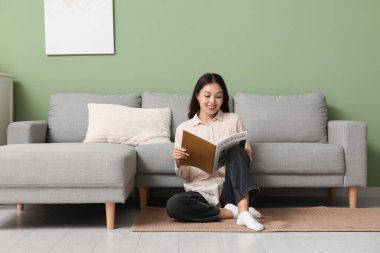 Genç Asyalı kadın evdeki kanepenin yanındaki halıda dergi okuyor.