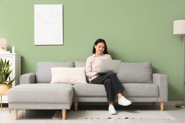 Genç Asyalı kadın evdeki gri koltukta dizüstü bilgisayar kullanıyor.
