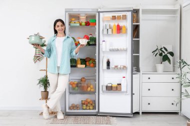 Mutfaktaki açık buzdolabının yanında tencere ve sebzelerle güzel bir kadın.