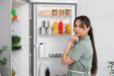 Mutfaktaki açık buzdolabının yanında düşünceli bir genç kadın.