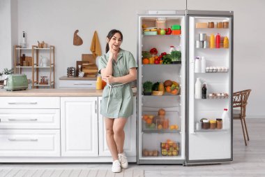 Mutfaktaki açık buzdolabının yanında düşünceli bir genç kadın.