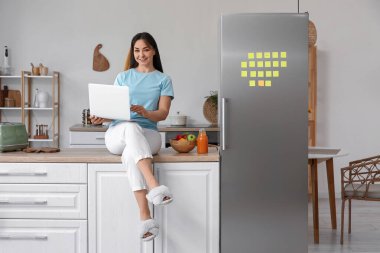 Bilgisayarlı güzel bir kadın mutfaktaki buzdolabının yanında oturuyor.