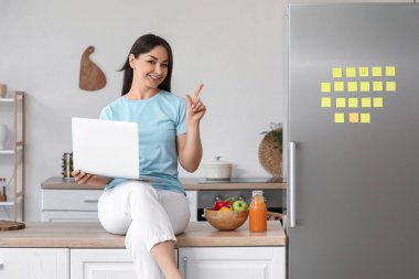 Bilgisayarlı güzel bir kadın mutfaktaki buzdolabını işaret ediyor.
