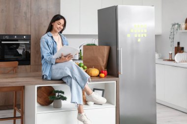 Mutfaktaki buzdolabının yanındaki masada dergi okuyan güzel bir kadın.