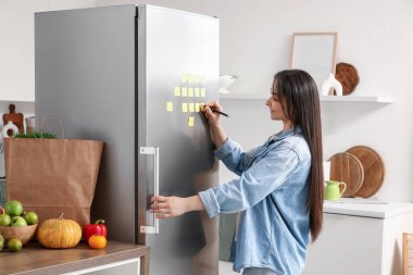 Güzel bir kadın mutfaktaki buzdolabına not yazıyor.
