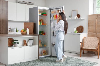 Mutfaktaki açık buzdolabının yanında düşünceli bir genç kadın.