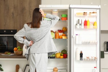 Mutfaktaki açık buzdolabının yanında düşünceli bir iş kadını.