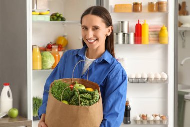 Mutfaktaki açık buzdolabının yanında yiyecek dolu bir çantası olan genç bir kadın.