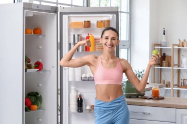 Elmalı genç bir kadın mutfaktaki açık buzdolabının yanında kaslarını gösteriyor.