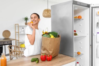 Mutfaktaki açık buzdolabının yanında yiyecek dolu bir çantası olan genç bir kadın.