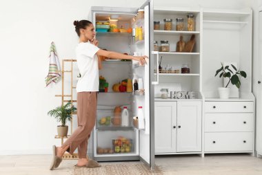 Mutfaktaki açık buzdolabının yanında düşünceli bir genç kadın.