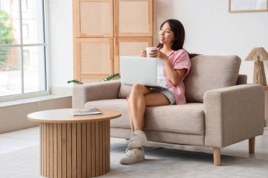 Evdeki kanepede oturan bir fincan kahve ve dizüstü bilgisayarlı genç Asyalı kadın.