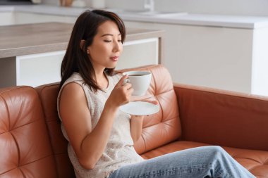 Mutfaktaki kanepede oturan bir fincan kahveli genç Asyalı kadın.